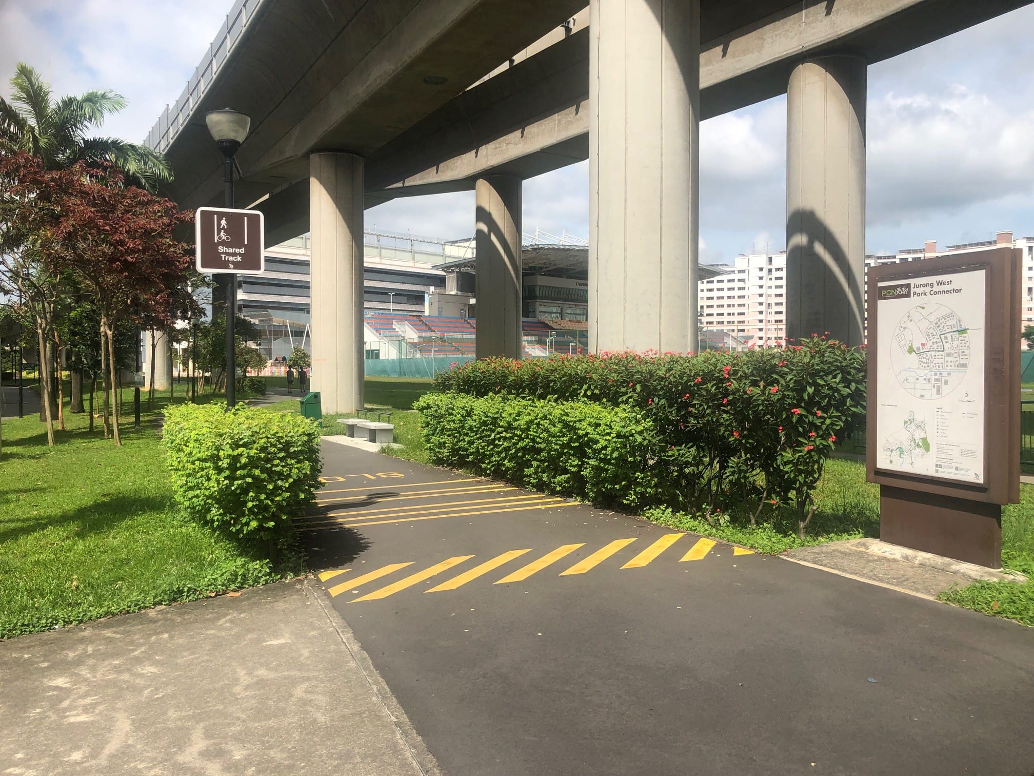 A shared track sign and map directory are near a walkway under a bridge surrounded by greenery.
